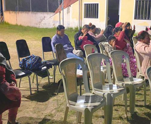 A group of people sit in chairs with eyes closed and hands raised