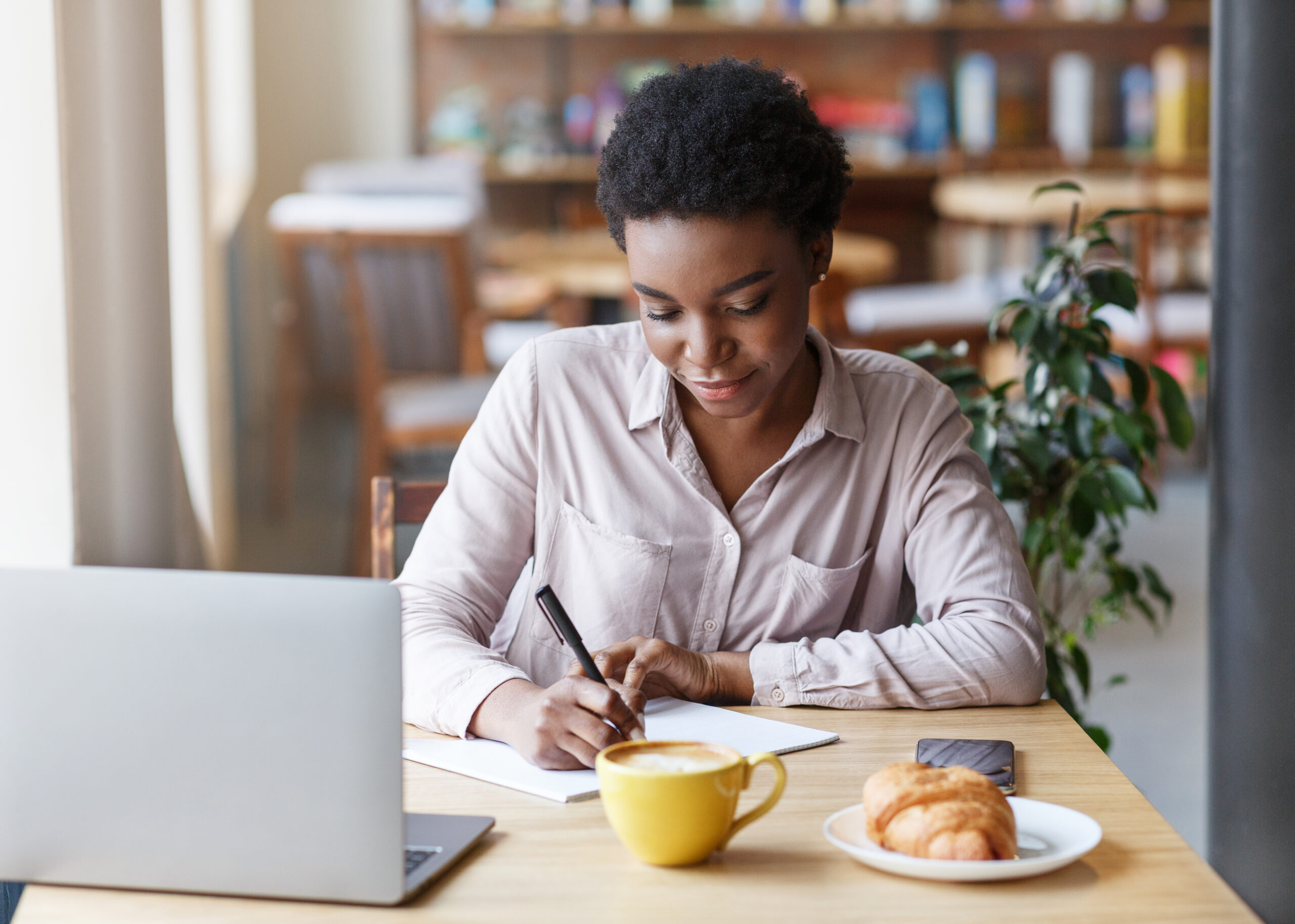 African American female student making notes in copybook while studying online at cafe